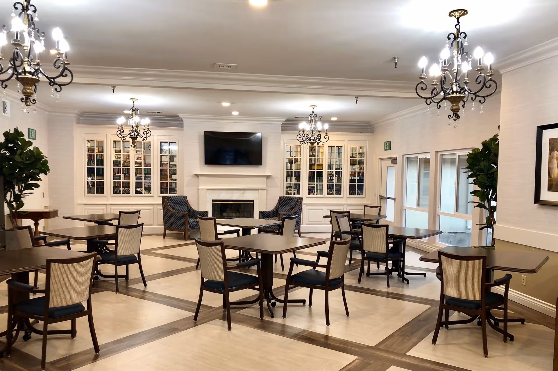 A spacious and well-lit common area with multiple tables and chairs arranged neatly. The room features elegant chandeliers hanging from the ceiling, a fireplace with a mounted flat-screen TV above it, and built-in glass-front cabinets filled with books. There are also two armchairs near the fireplace and large windows on one side letting in natural light. Potted plants and framed artwork decorate the walls.