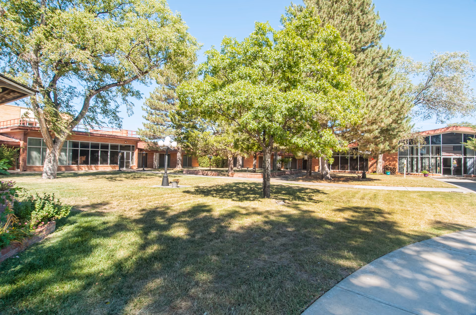 Outdoor view of a nursing home facility with a grassy lawn, several trees providing shade, a curved concrete walkway, and a brick building with large windows in the background under a clear blue sky.