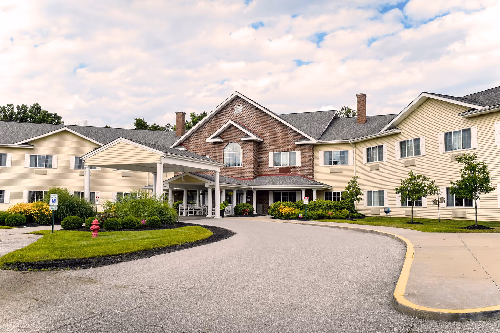 Front exterior view of Eden Vista Stow facility showing a large two-story building with beige siding and a central brick section. There is a covered entrance with white pillars, a driveway, landscaped greenery, and a fire hydrant in front.