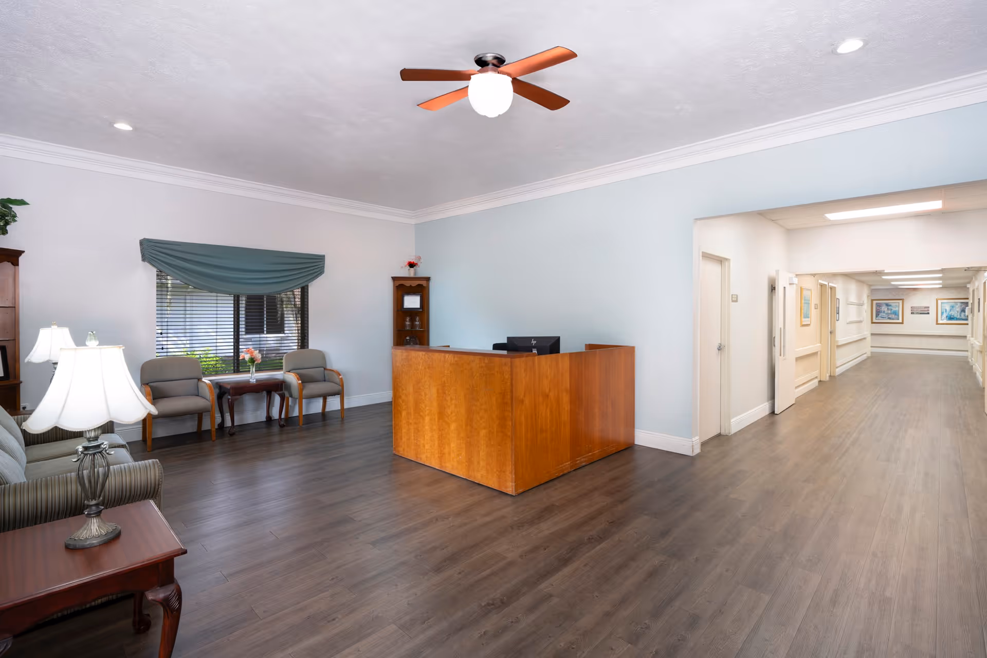 Reception area of Harborview Health Center Kissimmee with a wooden reception desk, two chairs and a small table with flowers near a window with blinds and a green valance. There is a ceiling fan with a light, a sofa with a lamp on a side table, and a hallway leading to other rooms.