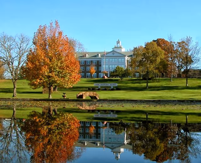 A large multi-story building with a cupola on top, surrounded by green lawns and trees with autumn foliage, reflected in a calm pond in the foreground. A stone bridge and a sign reading 'The Baptist Home' are visible on the lawn.