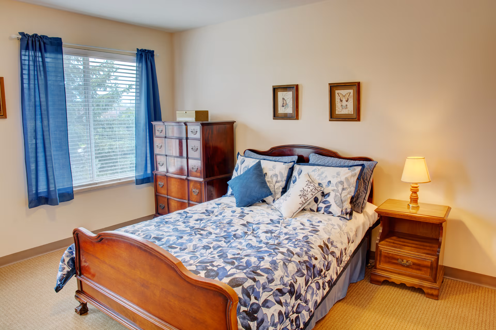 Bright bedroom featuring a wooden bed with blue floral bedding, a wooden dresser and nightstand with a lamp, plus a window with blue curtains.