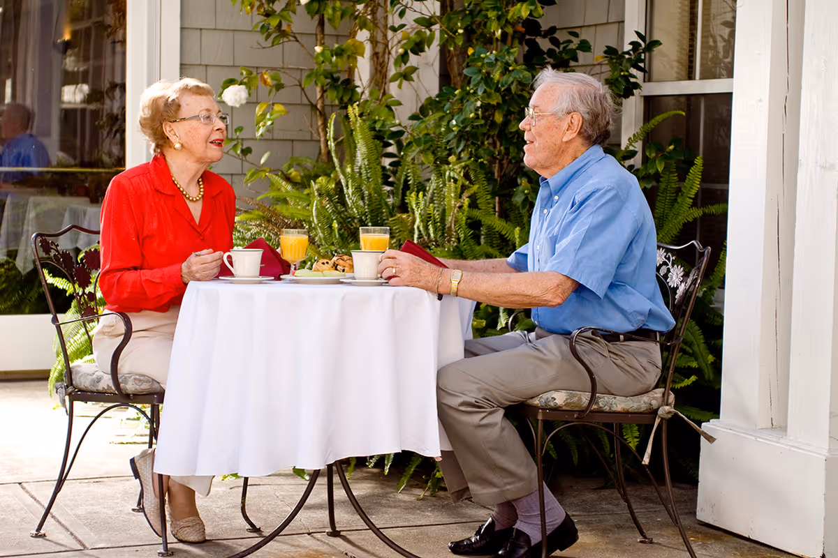 An elderly woman in a red blouse and an elderly man in a blue shirt sit at a small round table covered with a white tablecloth outdoors, enjoying coffee, orange juice, and pastries. They are engaged in conversation with greenery and a building wall in the background.