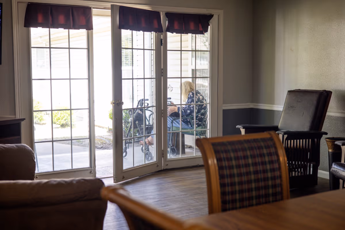 Interior common room with chairs and a table facing glass patio doors, and a person sitting outside next to a walker.