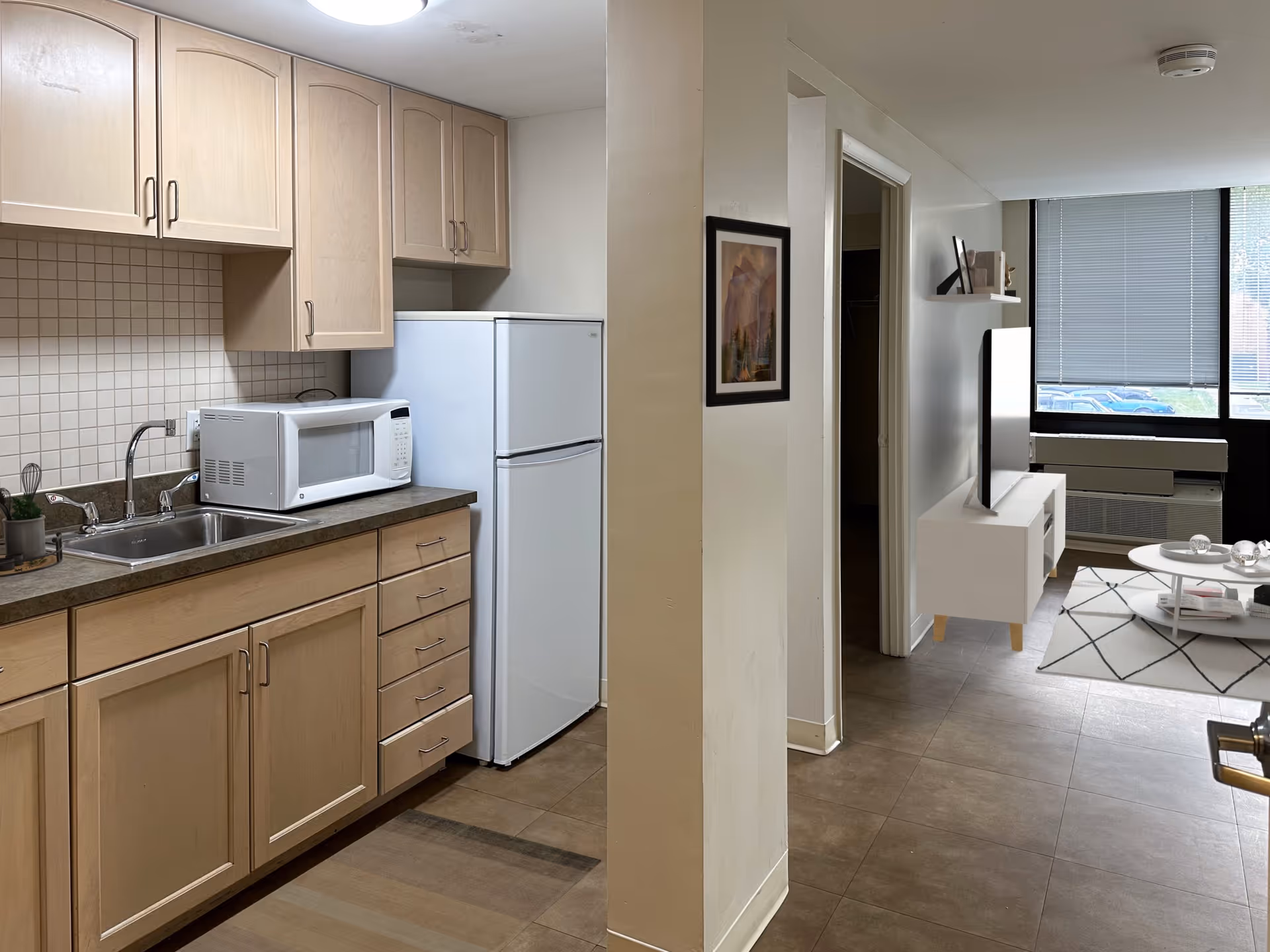 Interior view of a senior living facility apartment showing a small kitchen area with light wood cabinets, a microwave, a sink, and a white refrigerator. Adjacent to the kitchen is a hallway leading to a living room with a large window, a TV on a white stand, a coffee table, and a rug.