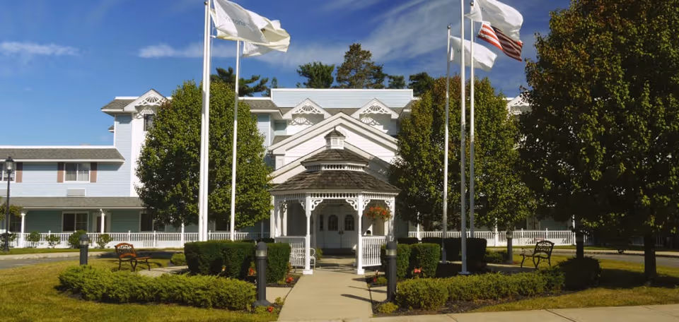 Front entrance of a white assisted living facility with a gazebo-style porte-cochere, flagpoles, benches, and landscaped trees and shrubs.