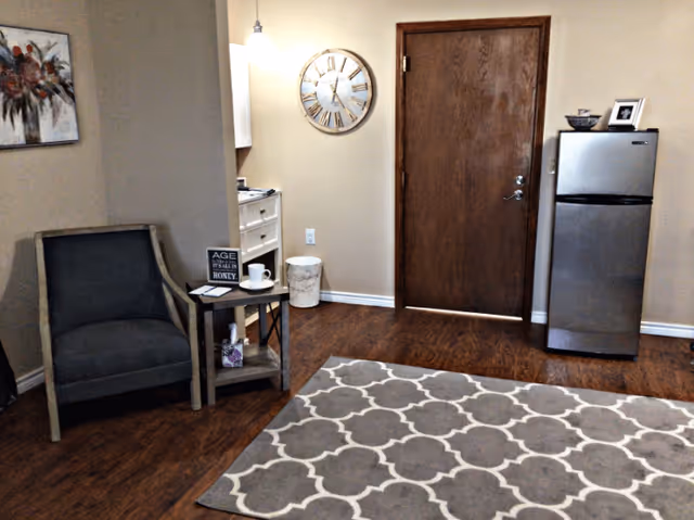 Interior room with a wooden door, a small stainless steel refrigerator topped with a bowl and framed photo, a large wall clock, a gray armchair next to a small side table with a cup, book, and decorative sign, a patterned area rug on wooden floor, and a piece of floral wall art.
