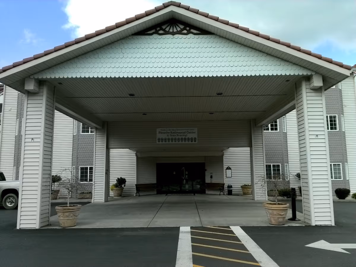 Covered entrance of a senior living facility with a peaked roof supported by white columns. There are potted plants on either side of the entrance and benches near the doors. The building exterior is light-colored with multiple windows.