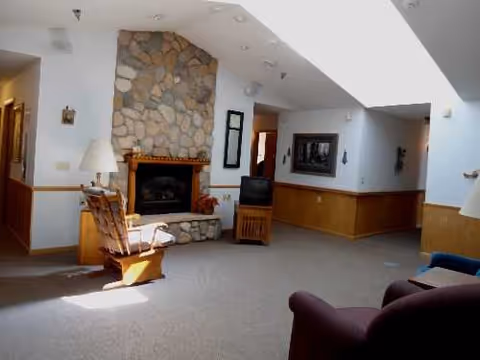 Cozy lounge area with stone fireplace, wooden chairs, and a TV in a senior living facility.