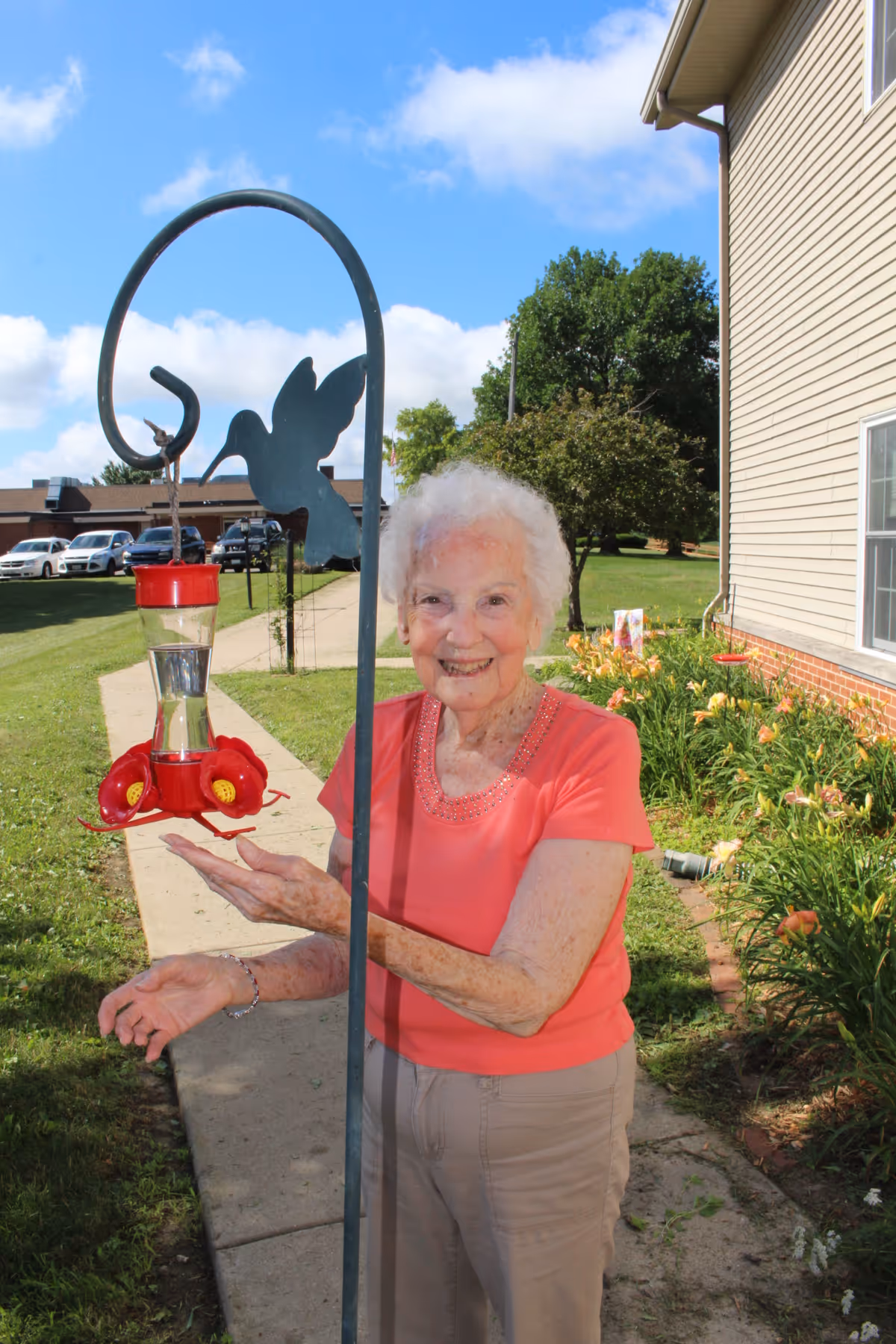 An elderly woman with white hair wearing a coral shirt and beige pants stands outside near a garden and a building. She is smiling and holding her hands near a red hummingbird feeder hanging from a metal pole shaped like a hummingbird. The background shows a sidewalk, grass, flowers, trees, and parked cars under a blue sky with some clouds.