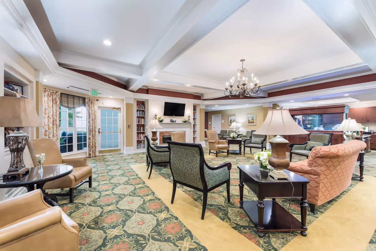 Spacious, well-lit senior living lounge with patterned carpet, multiple armchairs and sofas, side tables and lamps, and a TV above a fireplace.