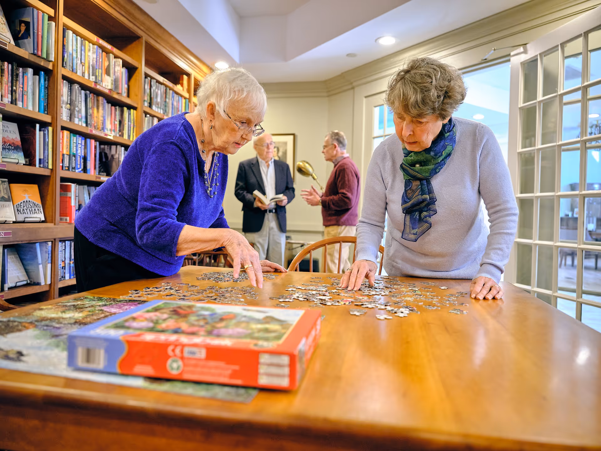 Two elderly women working together on a jigsaw puzzle at a wooden table in a room with bookshelves filled with books. In the background, two elderly men are standing and talking, one holding a book. The room has large windows and a cozy, well-lit atmosphere.
