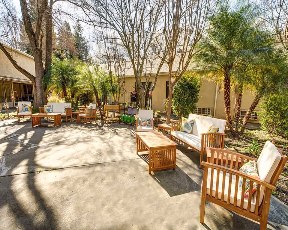 Sunny outdoor courtyard with wooden patio furniture and colorful cushions among trees and palms in front of a beige building.