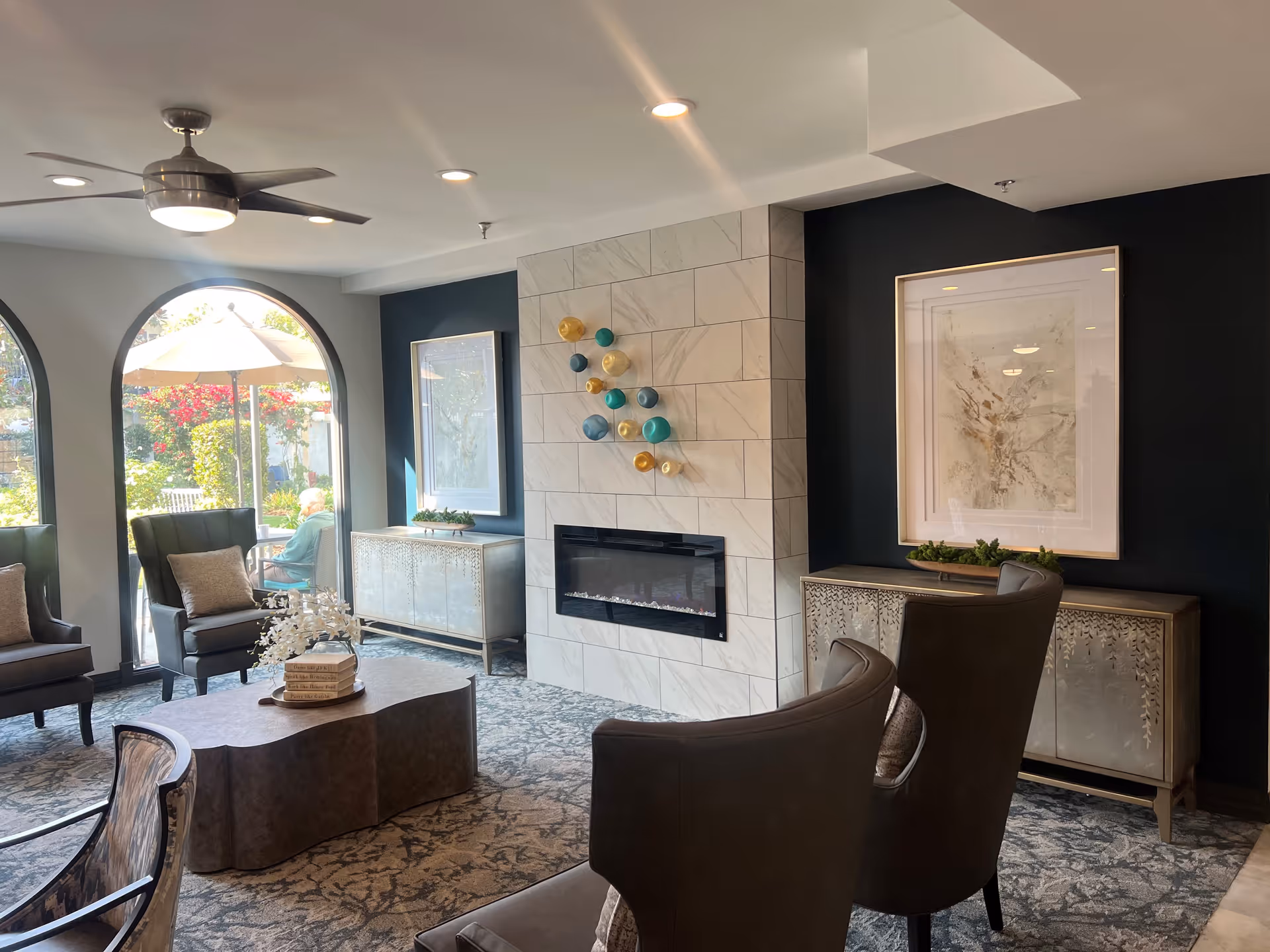 A cozy senior living facility common area with a modern fireplace set in a tiled wall. The room features comfortable leather chairs, a uniquely shaped coffee table with decorative books and flowers, and two cabinets with framed artwork above them. Large arched windows let in natural light and offer a view of an outdoor patio with an umbrella and greenery.