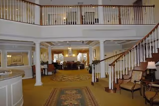 Spacious interior common area with a staircase on the right, carpeted floors, columns, and seating areas with chairs and tables. The upper level has a railing overlooking the lower level, and the space is well-lit with lamps and ceiling lights.