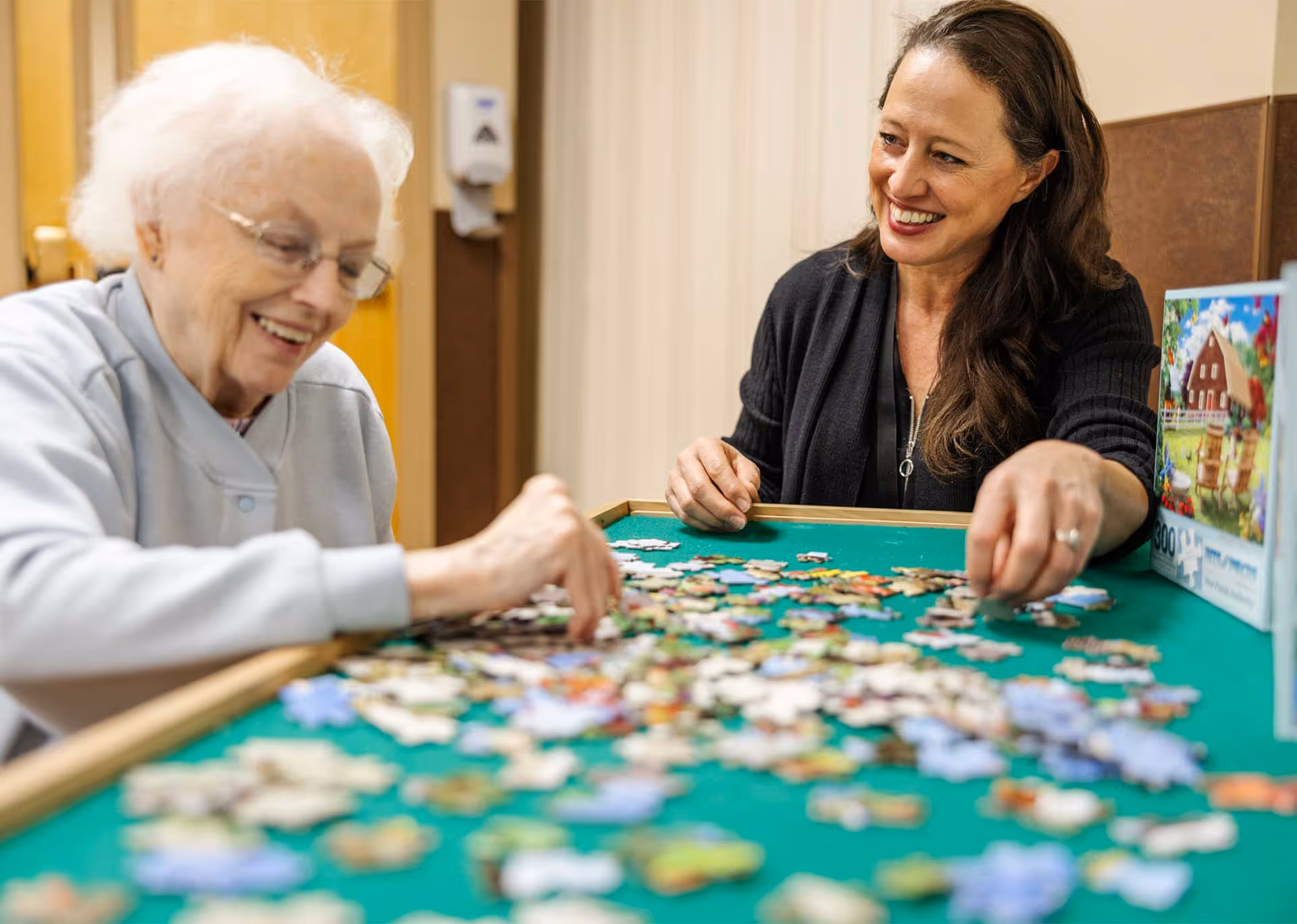 An elderly woman and a younger woman sitting at a table working on a jigsaw puzzle together, both smiling and engaged in the activity.