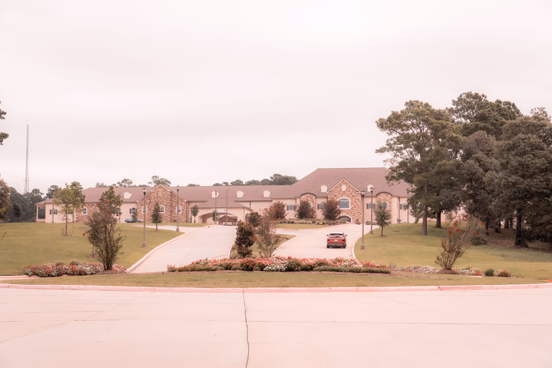 Front exterior view of Worthington Manor, a large building with a stone facade and multiple windows, surrounded by green lawns, trees, and flower beds. A driveway leads up to the entrance with a single car parked on it.