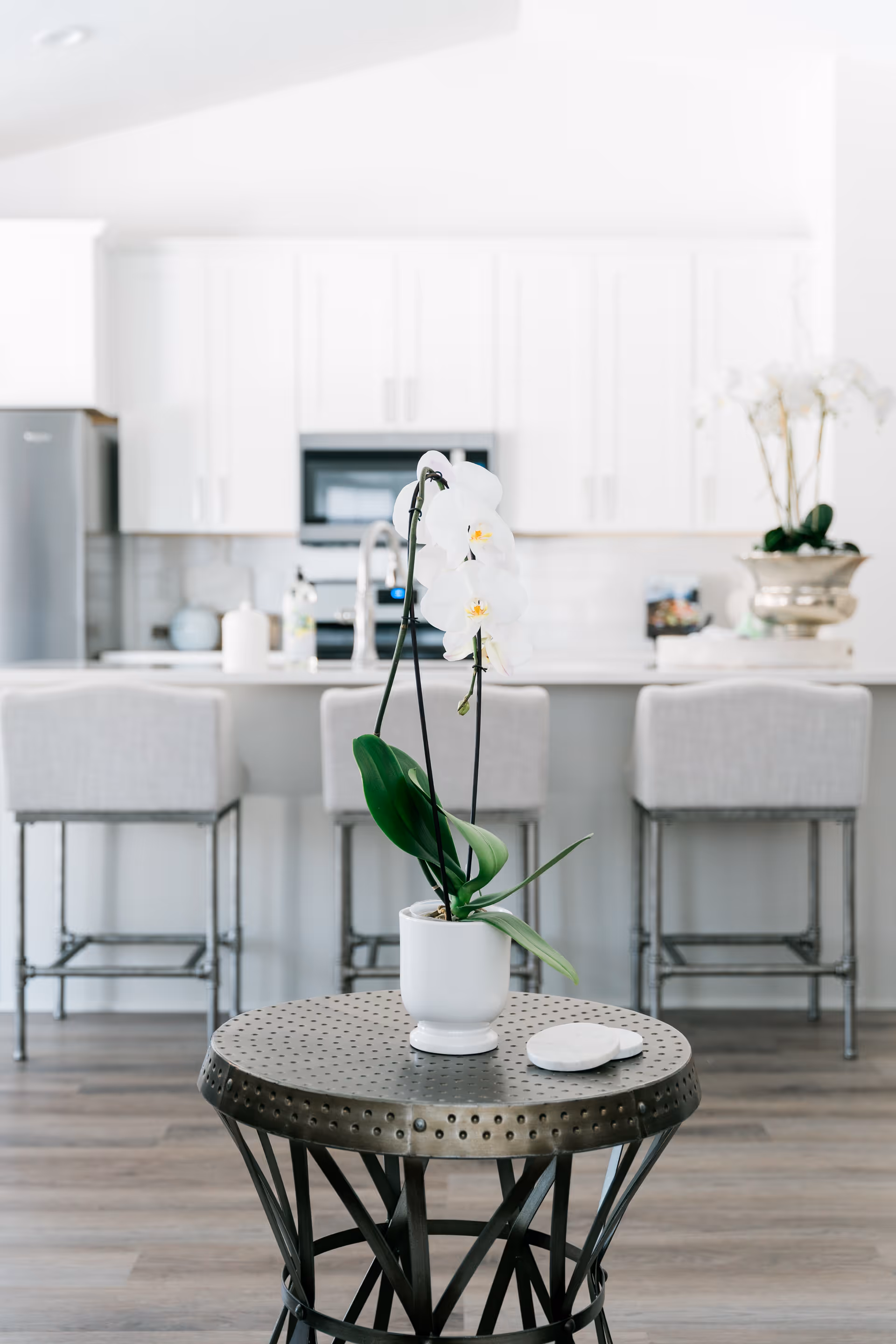 Small metal side table topped with a white potted orchid in front of a bright modern kitchen island with three upholstered bar stools.