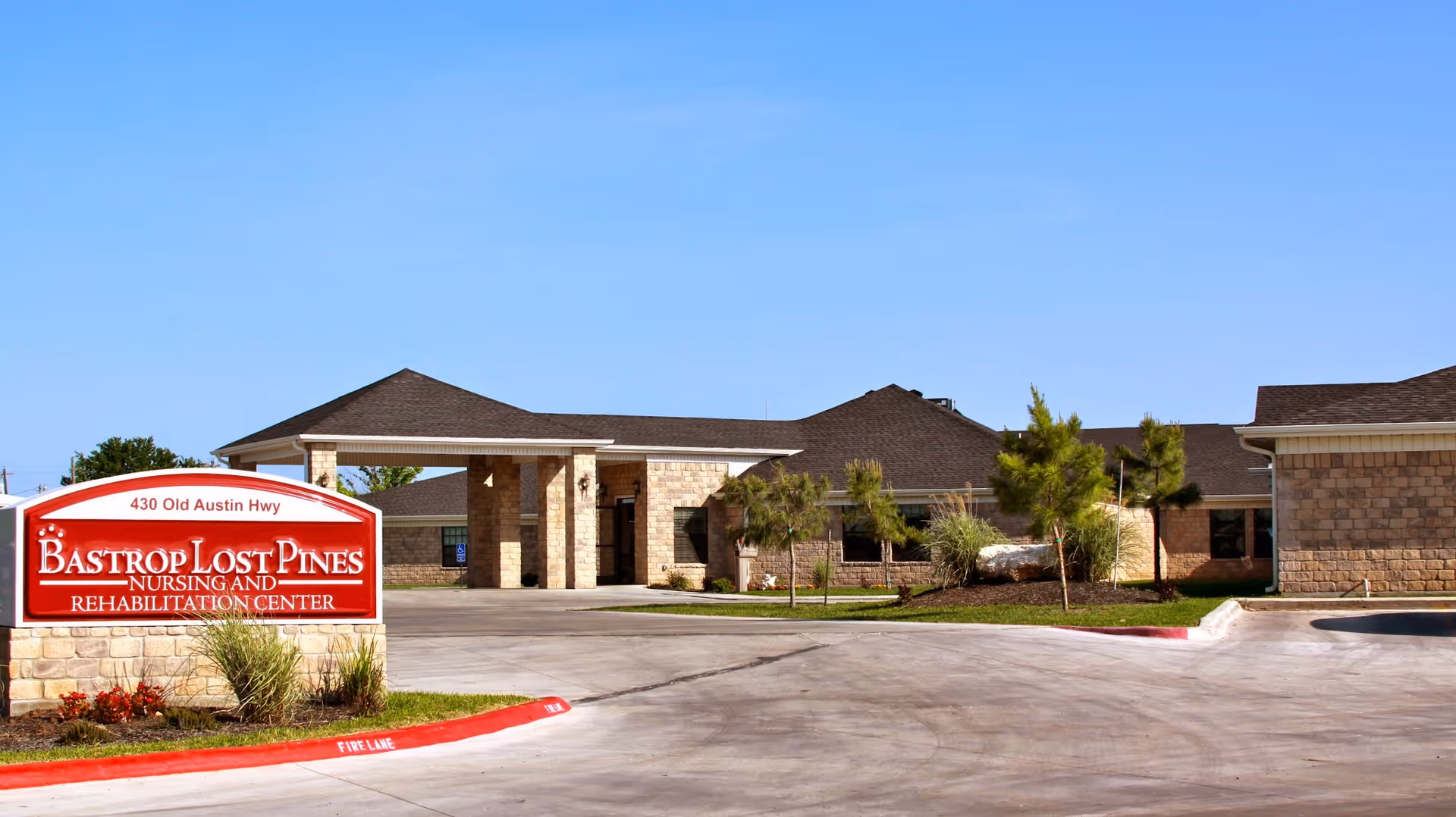 Front entrance of Bastrop Lost Pines Nursing and Rehabilitation Center with a red sign, driveway, and low landscaping.