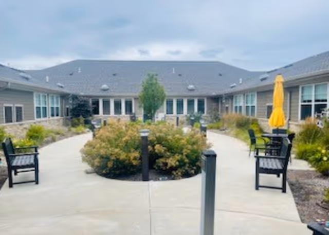 Outdoor courtyard area at a senior living facility with a paved walkway, benches, bushes, and a yellow umbrella. The courtyard is surrounded by single-story buildings with multiple windows under a cloudy sky.