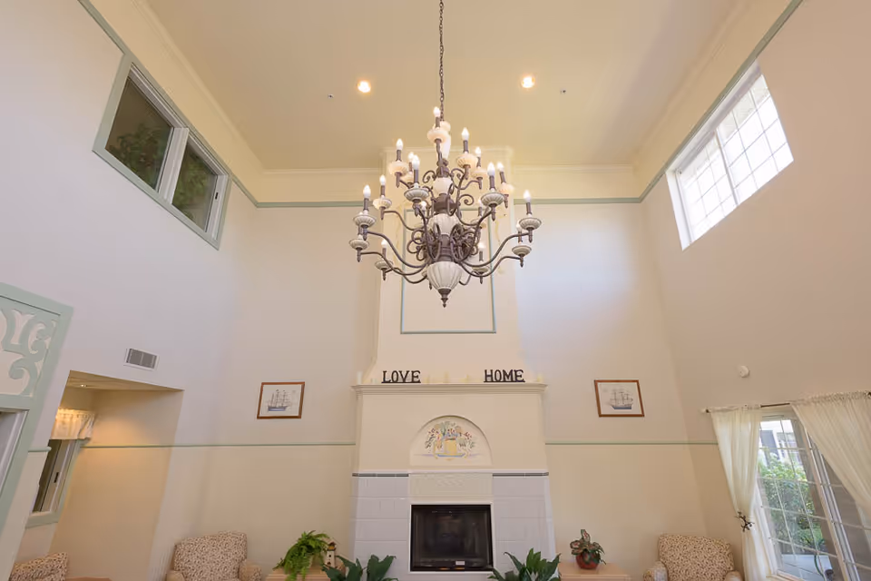 A spacious living room with high ceilings featuring a large ornate chandelier hanging in the center. The room has a fireplace with decorative tiles and the words 'LOVE' and 'HOME' displayed on the mantel. There are framed pictures on the walls, several plants, and comfortable armchairs. Large windows allow natural light to fill the space.
