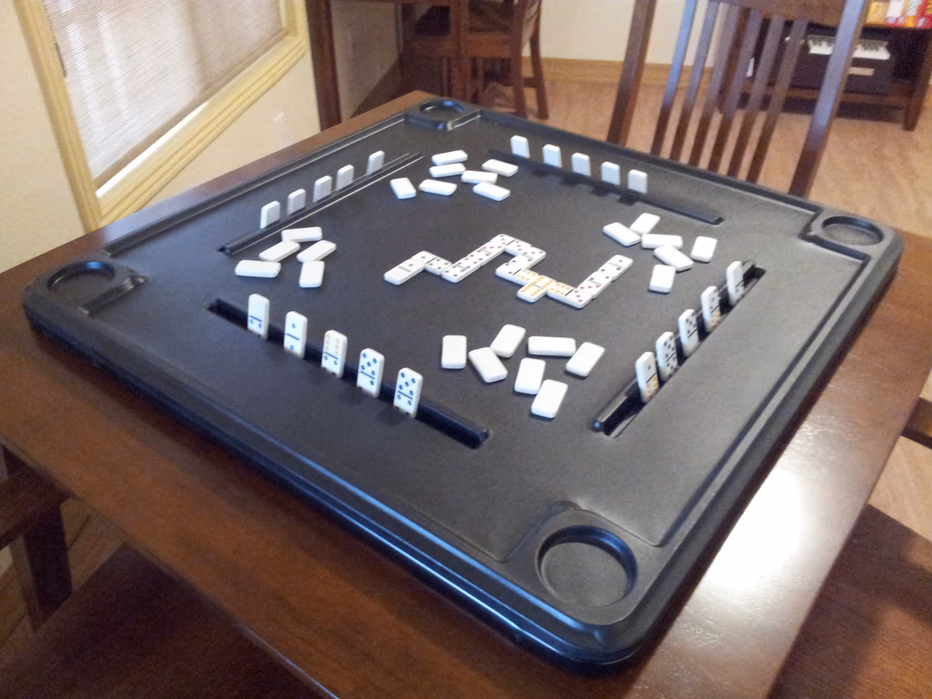 A black square game board with domino tiles arranged on it, placed on a wooden table in a room with wooden chairs and a window with blinds.