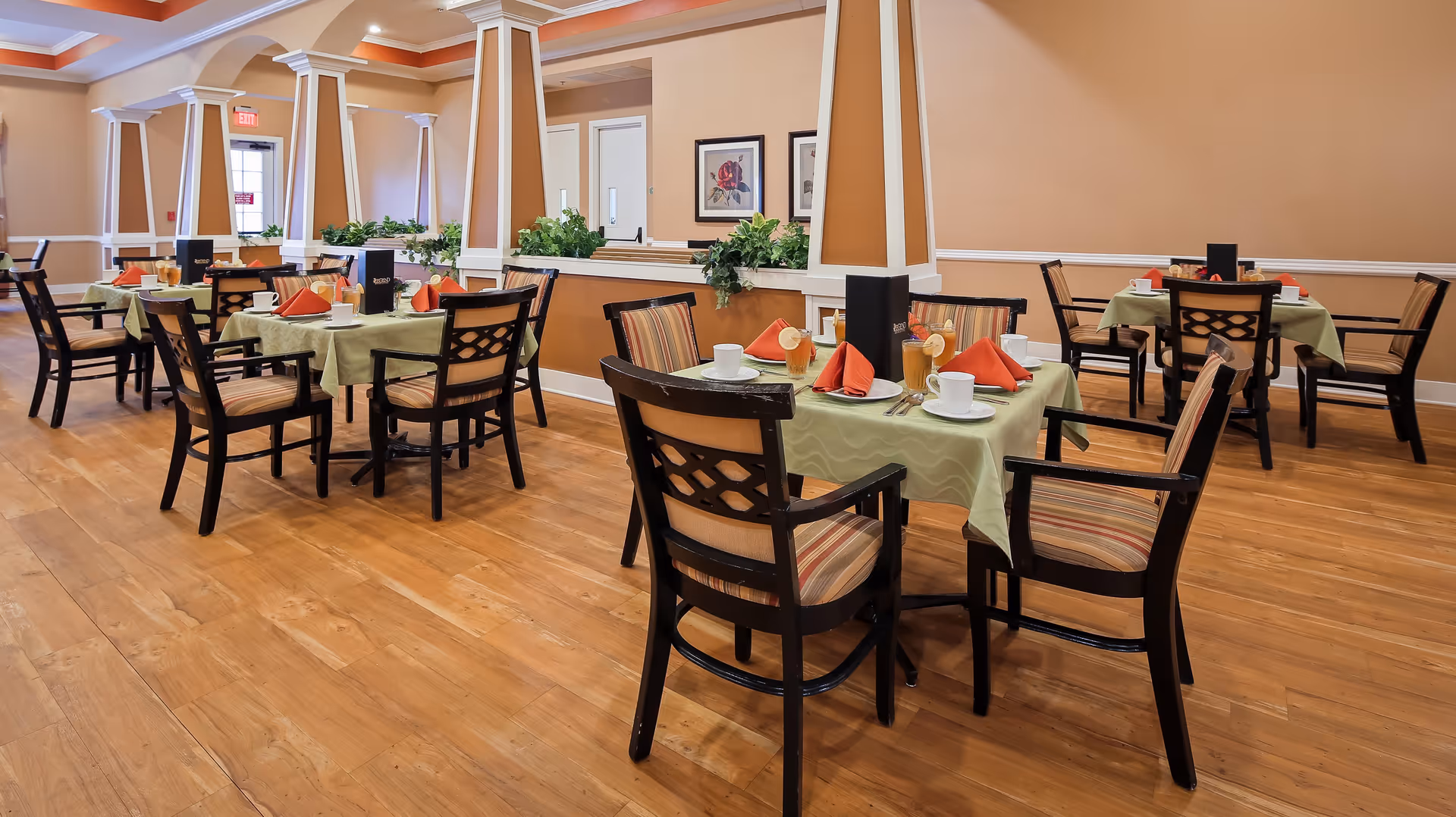 A dining room in an assisted living facility with several tables set with green tablecloths, orange folded napkins, white cups, and glasses. The room features wooden floors, beige walls, decorative columns, and framed artwork on the walls. Chairs with striped cushions surround the tables.