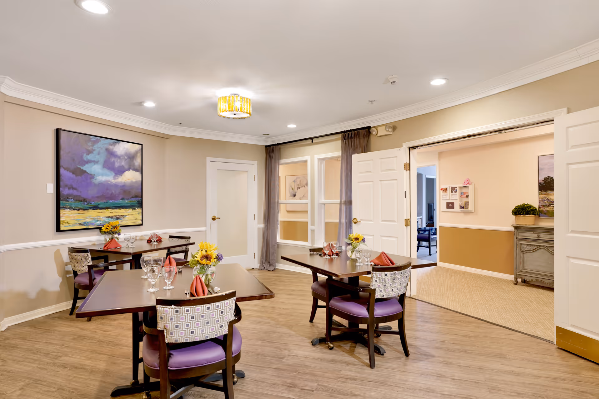 A dining room in a senior living facility with three wooden tables, each set with wine glasses, napkins, and small flower arrangements. The room has beige walls, a wooden floor, a colorful painting on one wall, and double doors leading to another room.