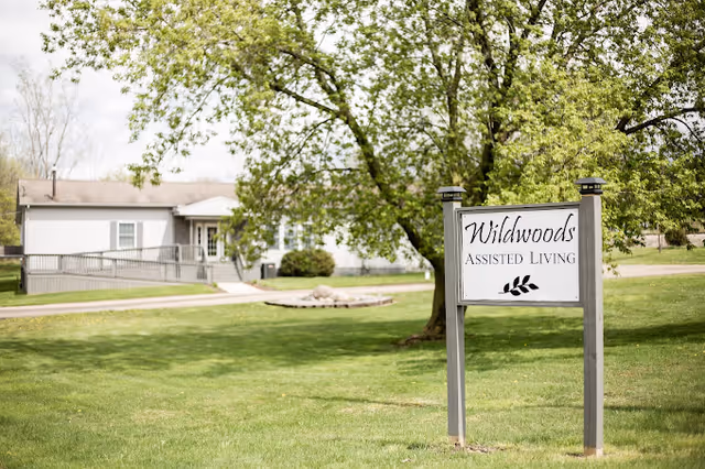 A sign reading 'Wildwoods Assisted Living' stands on a grassy lawn with a large tree nearby. In the background, there is a single-story building with a ramp leading to the entrance, surrounded by green grass and trees.