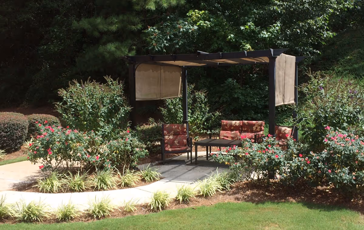 Outdoor seating area with a pergola structure providing partial shade over cushioned chairs and a table, surrounded by green bushes and flowering plants in a garden setting.