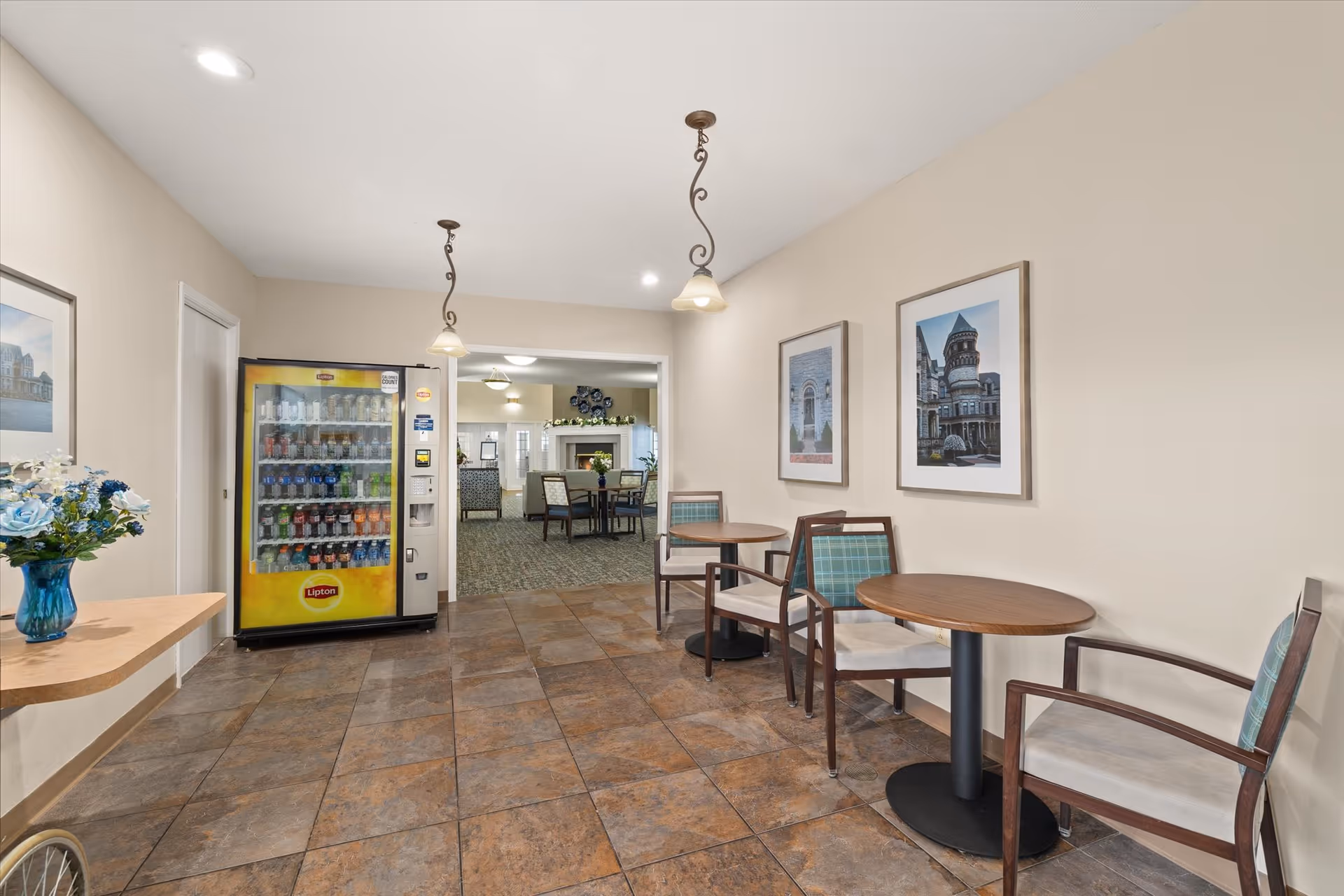 A seating area in a senior living facility with two small round tables and four chairs with green plaid cushions. There is a yellow Lipton vending machine stocked with drinks on the left side. The floor is tiled with brown and gray tiles, and the walls are beige with two framed pictures. Two pendant lights hang from the ceiling, and through an open doorway, a lounge area with more seating and a fireplace is visible.