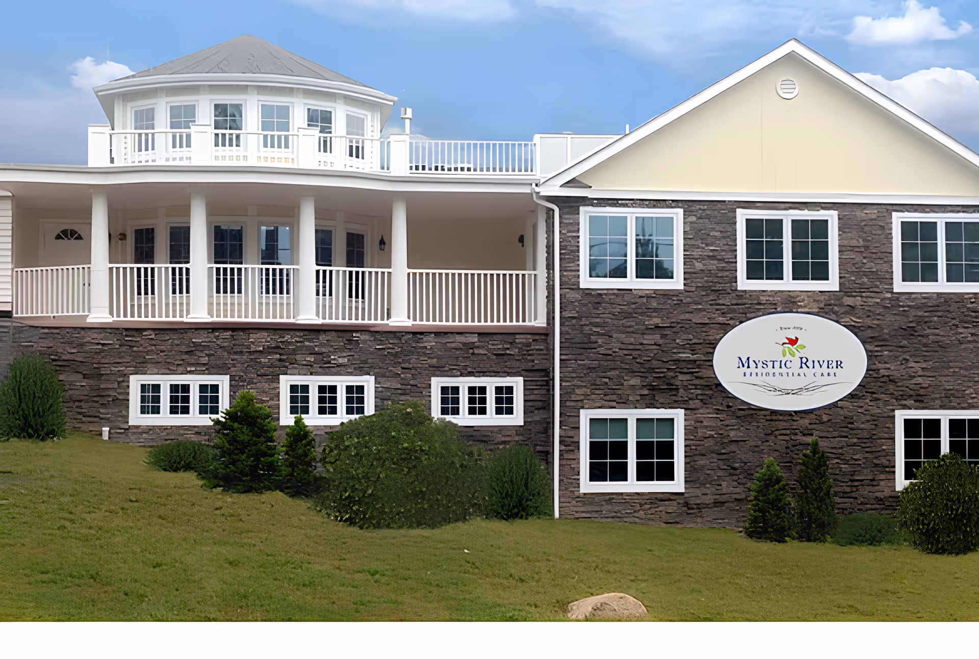 Exterior view of Mystic River Residential Care building featuring a stone facade with multiple windows, a white balcony with columns, and a sign displaying the facility's name. The building is surrounded by green grass and shrubs under a partly cloudy sky.