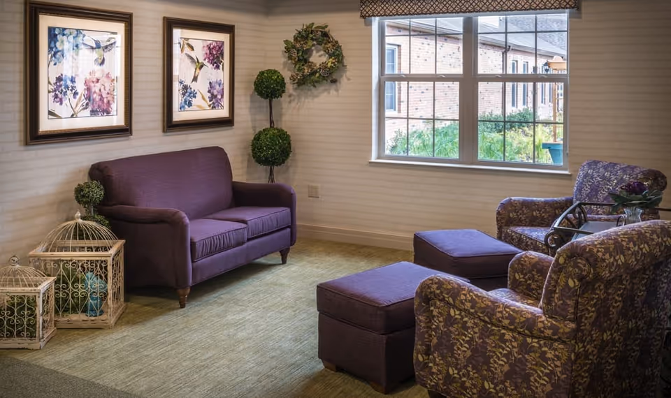 Cozy seating area with a purple loveseat, patterned armchairs and ottomans beside a window and decorative wall art.