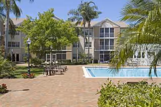 Outdoor area of a senior living facility named Coral Oaks featuring a swimming pool, lounge chairs, palm trees, and a multi-story building in the background under a clear sky.