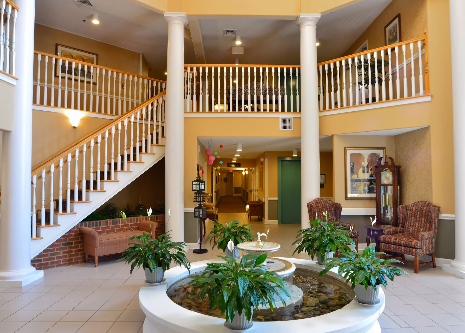 Interior view of a senior living facility lobby with a central water fountain surrounded by potted plants. There are two white columns supporting a balcony with white railings. A staircase with white balusters and wooden handrails leads to the upper floor. The lobby has comfortable seating including two patterned armchairs and a small sofa. A grandfather clock and framed artwork are visible on the walls. The space is well-lit with ceiling lights and has a warm, welcoming atmosphere.