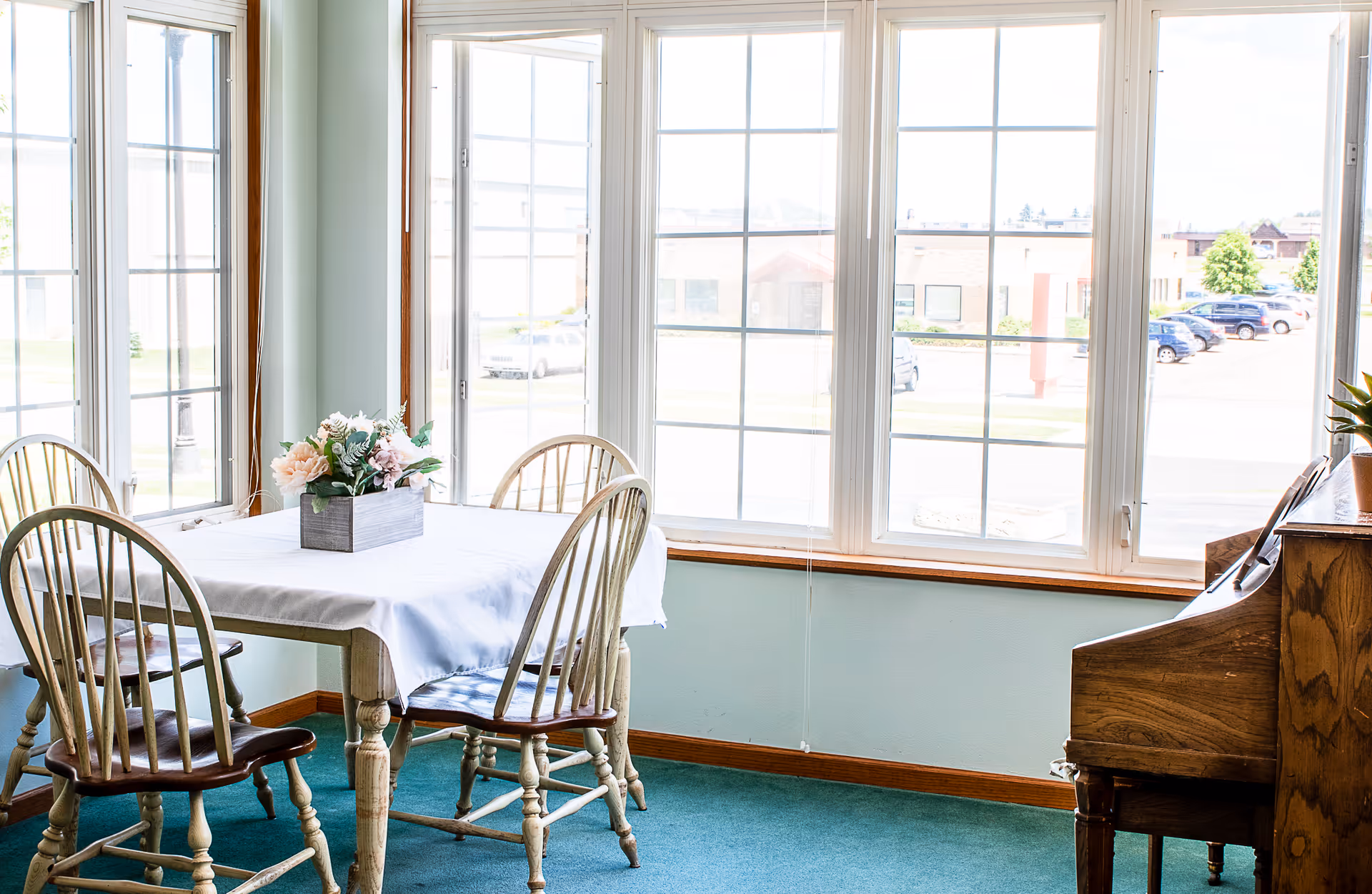 A bright dining area with large windows letting in natural light. There is a wooden table covered with a white tablecloth and four wooden chairs around it. A small floral arrangement in a rectangular vase is placed on the table. To the right, there is a wooden piano with a small plant on top. Outside the windows, a parking lot and some buildings are visible.