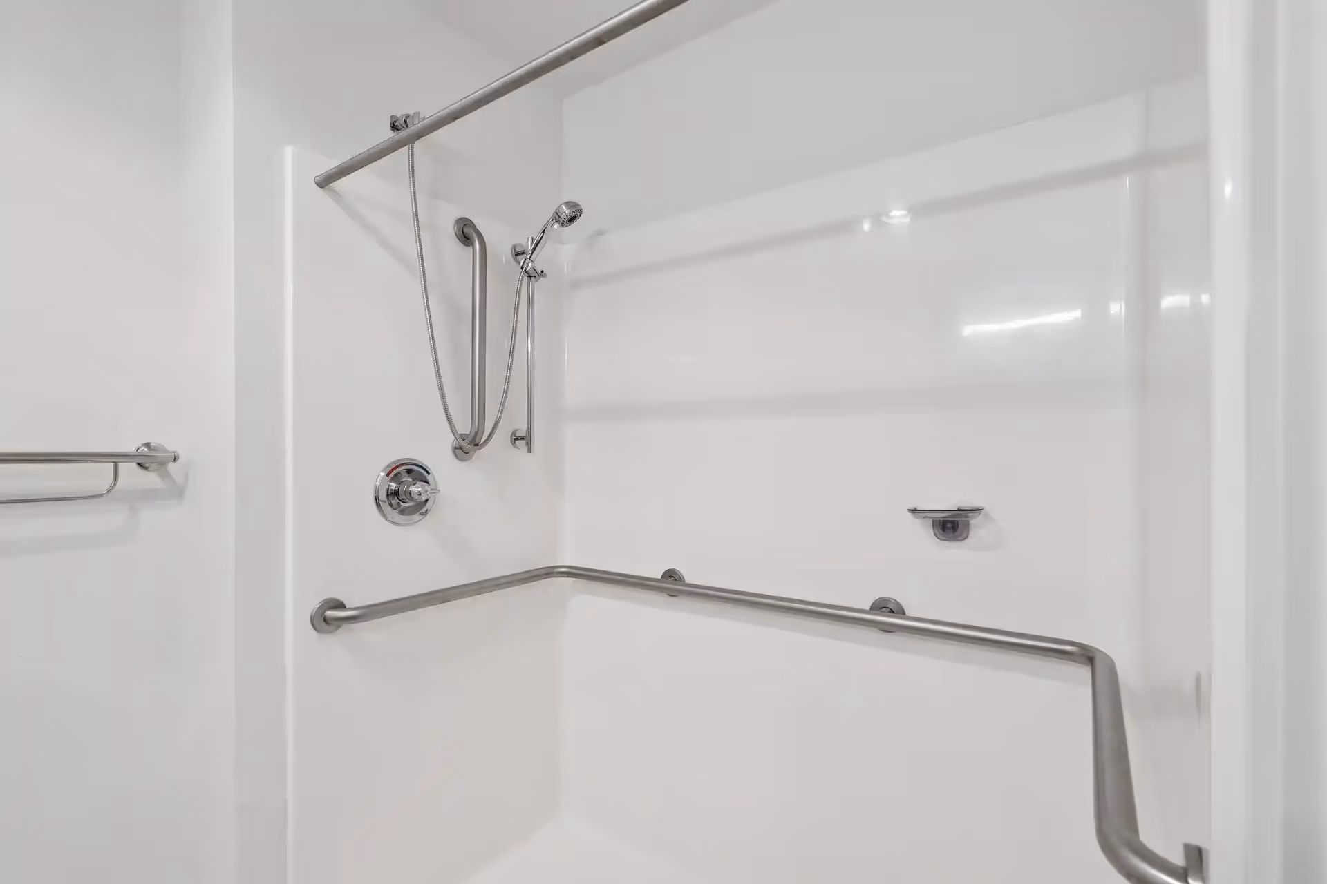 A white shower area with stainless steel grab bars on the walls and a handheld showerhead attached to a vertical grab bar. There is also a towel bar mounted on the adjacent wall.