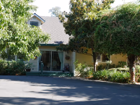 Front exterior view of a single-story residential building with a sloped roof, surrounded by trees and greenery. There is a paved driveway or parking area in front of the building, which has a sliding glass door and windows partially covered by curtains.
