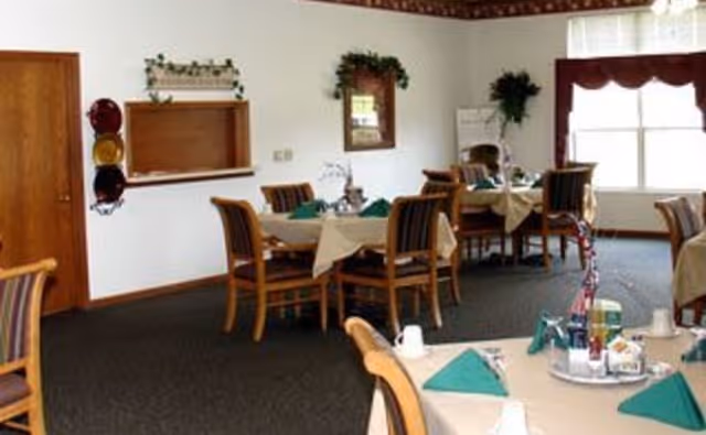 Dining room with several tables covered with beige tablecloths and green napkins, wooden chairs with striped cushions, a window with a valance, and wall decorations including a mirror and a plant.