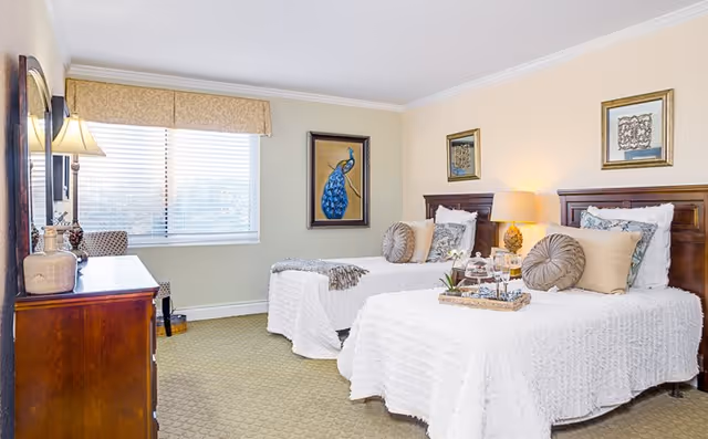A bright and cozy bedroom with two twin beds covered in white textured bedspreads, each adorned with decorative pillows. A wooden dresser with a lamp and decor items is positioned against one wall, and a window with blinds and a valance lets in natural light. The walls are decorated with framed artwork, including a painting of a blue peacock.