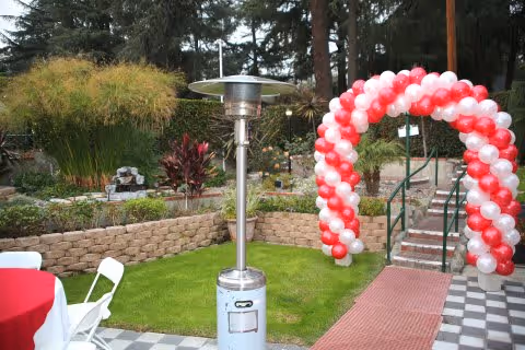Outdoor garden area with a red and white balloon arch over a stairway, a tall outdoor heater, green grass, stone retaining walls, and various plants and trees in the background.