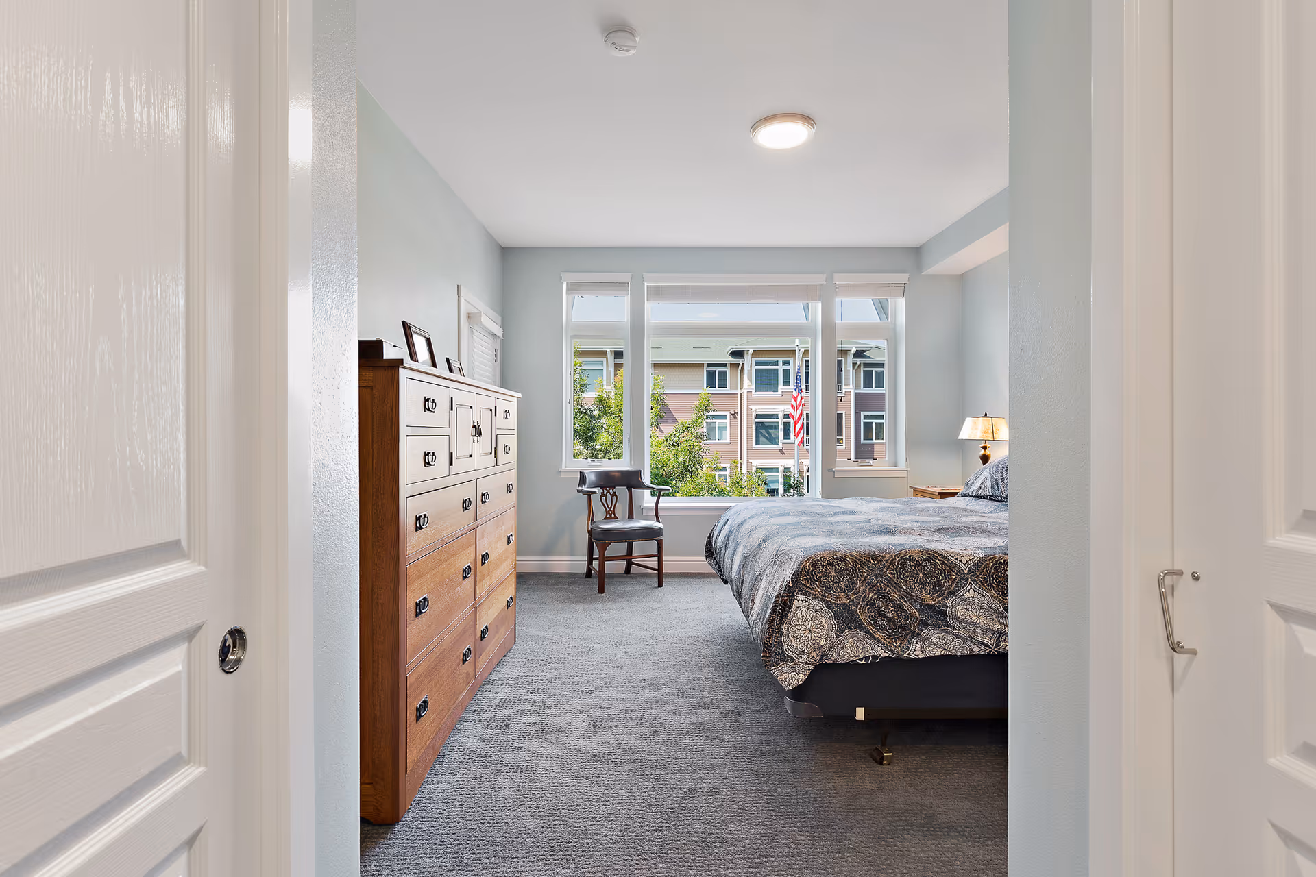 View into a bedroom with a large window showing an outside building and an American flag. The room has a bed with patterned bedding, a wooden dresser with framed photos on top, a wooden chair near the window, and a bedside table with a lamp. The walls are painted light blue and the floor is carpeted.