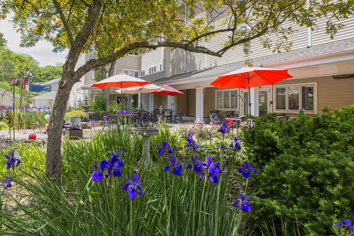 A landscaped courtyard with purple irises, patio tables and red umbrellas in front of a multi-story senior living building.