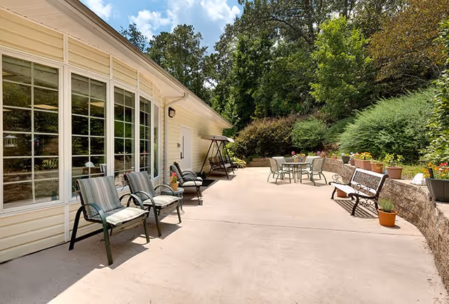 Outdoor patio area at Lakeview Estates - Memory Care Community with several chairs, a bench, a table with chairs, potted plants, and greenery surrounding the space under a partly cloudy sky.