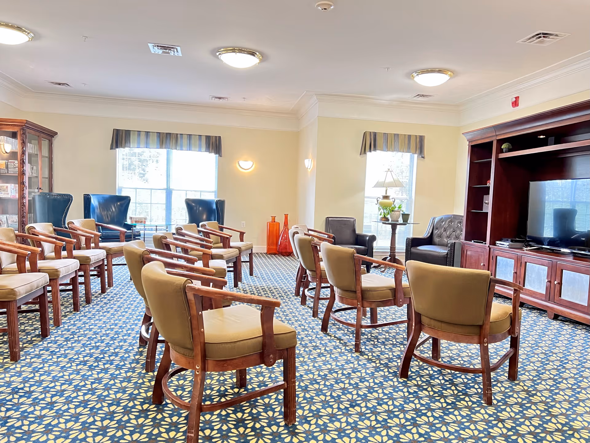 A well-lit common room with multiple wooden chairs arranged in rows facing a large wooden entertainment center with a flat-screen TV. The room has patterned blue and yellow carpet, two large windows with striped valances, and additional seating including two blue armchairs and two black leather chairs near the windows. There are decorative items such as a lamp on a small table and large orange vases in the corner.