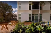 Outdoor garden area with raised planting beds filled with green leafy vegetables in front of a modern white building with large windows. There is a wooden picnic table on a paved area to the left and trees in the background under a partly cloudy sky.