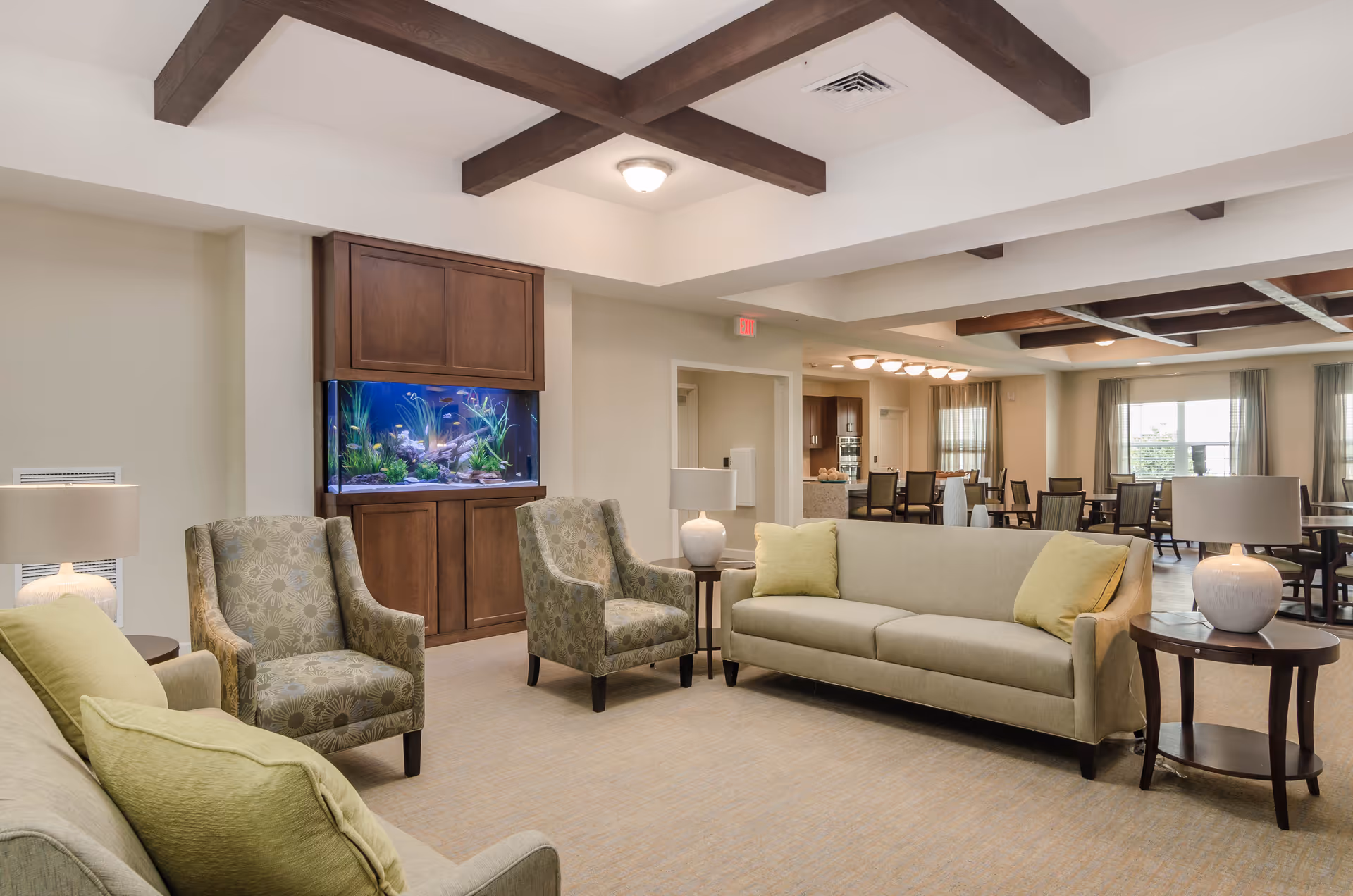 Bright senior living lounge with sofas, armchairs, a built-in aquarium, and dining tables in the background.