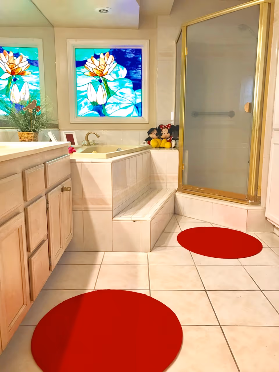 A bathroom with a tiled floor and walls, featuring a built-in bathtub with a step, a glass-enclosed shower with a gold frame, a wooden vanity with cabinets, two red circular rugs on the floor, a stained glass window with floral design, and two plush toys of Mickey and Minnie Mouse placed on the bathtub edge.