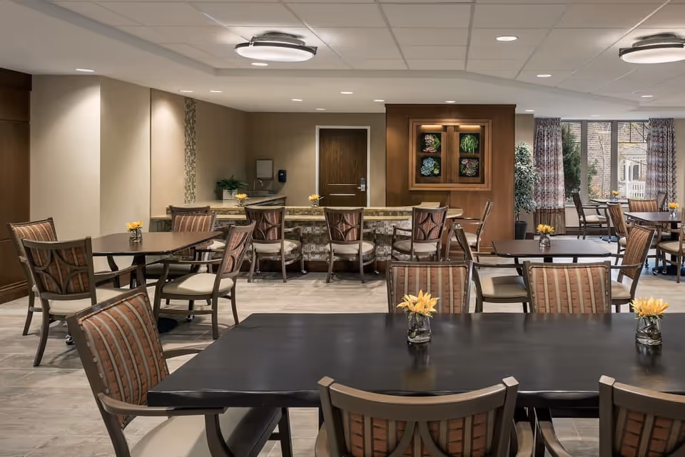 A spacious dining room with multiple dark wood tables and cushioned chairs arranged neatly. Small vases with yellow flowers are placed on each table. The room features a tiled floor, beige walls, a ceiling with recessed lighting, and large windows with patterned curtains letting in natural light. There is a wooden cabinet with decorative plants on the far wall and a door in the background.