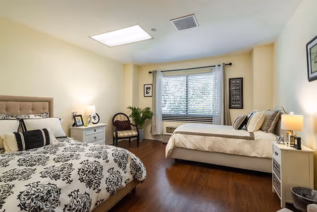 A well-lit bedroom with two beds, one with a black and white patterned comforter and the other with a beige comforter. The room has wooden flooring, a window with white curtains, a small white nightstand with a lamp and picture frame, a chair with a cushion, and a potted plant near the window.